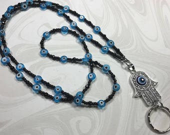 May include: A beaded lanyard featuring blue evil eye beads and black seed beads. The lanyard includes a silver-tone Hamsa hand charm with a central evil eye design and a key ring. The beads are approximately 0.3 inches in diameter.