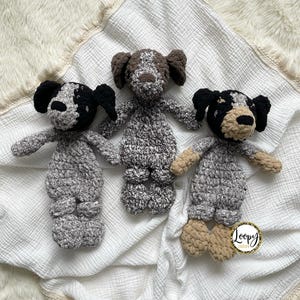 May include: Three crocheted dog plushies in black, brown, and white on a white crocheted blanket with a white fringe. The blanket has a small tag that says "Loopy Couture".