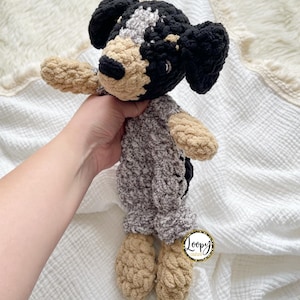 May include: A handmade crochet dog plushie in black, tan, gray, and white. The dog has floppy black ears, a tan snout, and a gray and white patch on its head. The body is gray, and the paws are tan. The plushie is soft and cuddly.