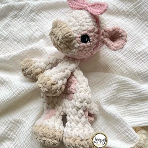May include: A crocheted stuffed animal cow with pink ears and a pink bow. The cow has white and brown fur and black eyes. The cow is lying on a white blanket with a fringe border. The blanket has a small tag that says 'Loopy Loofah'.