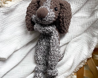 May include: A handmade crochet dog toy in shades of brown and white. The dog has floppy ears, a white patch on its head, and a textured body. The toy is lying on a white, textured blanket, with a wooden basket in the background.