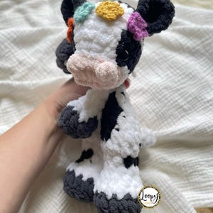 May include: A crocheted stuffed cow toy with black and white spots. The cow has a pink nose and black eyes. The cow has colorful crocheted flowers on its head. The cow is standing on a white blanket. The text 'Loopy Creations' is visible on a tag attached to the cow's leg.