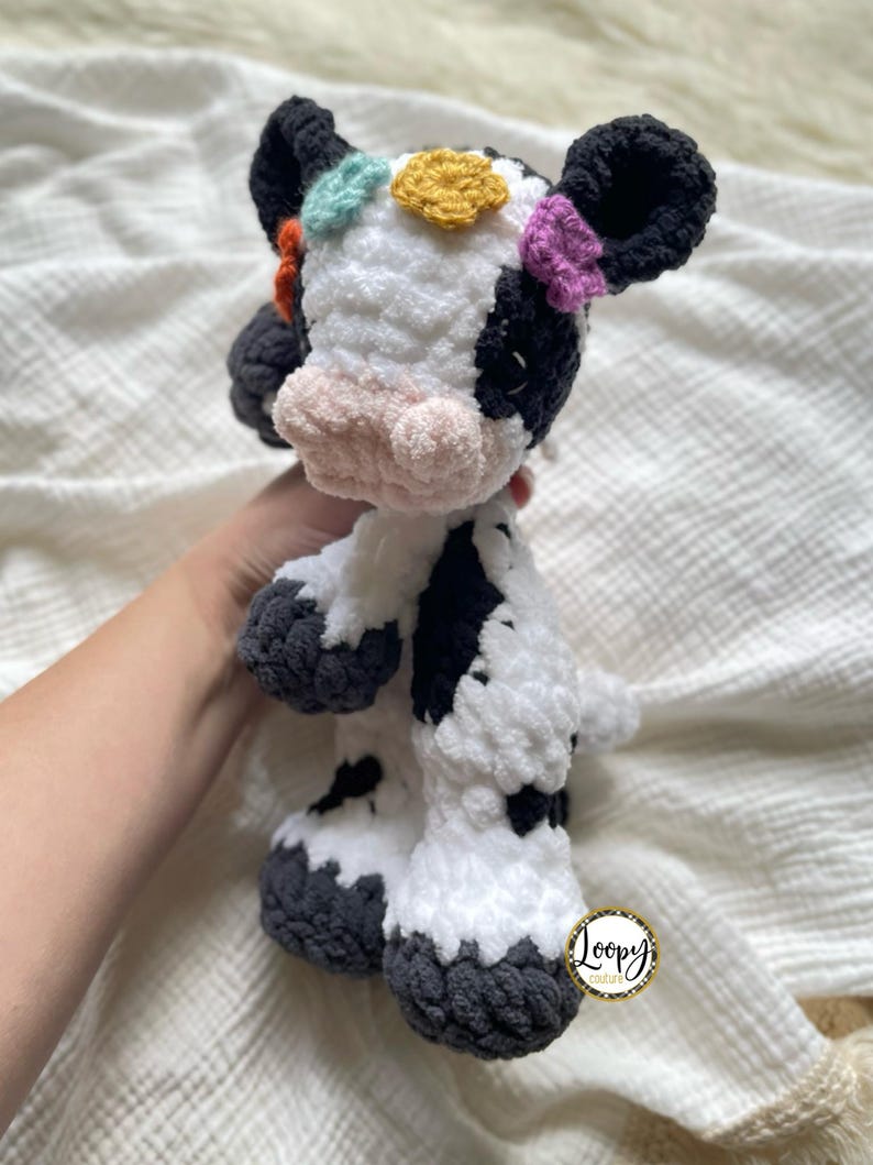 May include: A crocheted stuffed cow toy with black and white spots. The cow has a pink nose and black eyes. The cow has colorful crocheted flowers on its head. The cow is standing on a white blanket. The text 'Loopy Creations' is visible on a tag attached to the cow's leg.