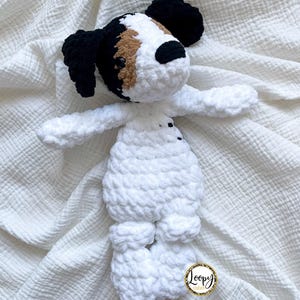 May include: A handmade crochet dog toy in white, black, and brown. The dog has floppy ears, a white body, and outstretched arms and legs. The toy is made of soft, textured yarn and is designed to be cuddly.