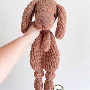 May include: A brown crocheted dog toy with a knotted tail and legs. The toy has a pink nose and black eyes.