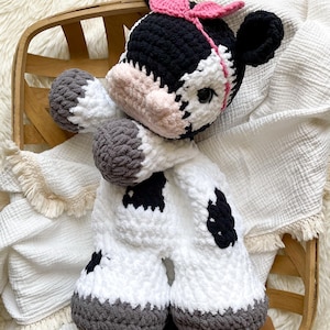 May include: A handmade crochet cow plushie with a black and white body, gray hooves, and a pink bow. The cow has black ears and spots, and a light pink muzzle. The plushie is in a wooden basket on a white blanket.