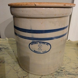 May include: A beige ceramic crock with a brown lid, featuring two horizontal blue stripes and a blue oval stamp that reads Marshall Pottery, Inc. 2 Marshall, Texas. The cylindrical container has a smooth, glazed finish and a wide rim.