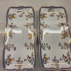 Peut inclure: Deux plateaux de service rectangulaires en porcelaine à bords festonnés, présentant une base blanche ornée de motifs floraux colorés en jaune, rose et vert, rehaussés d'une bordure dorée et d'une bordure bleue foncée.