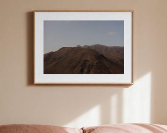 May include: A rectangular landscape photograph in a light-toned wooden frame featuring a mountain range under a pale sky. The image displays dark brown, rugged mountain slopes with visible ridges and shadows, contrasted against a soft, light blue sky.