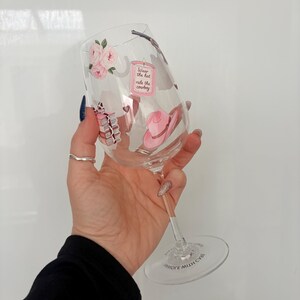 May include: A clear wine glass with a stem, decorated with pink roses, a pink cowboy hat, and the text "Wear the hat ride the cowboy". The glass has a wide bowl and a clear base.