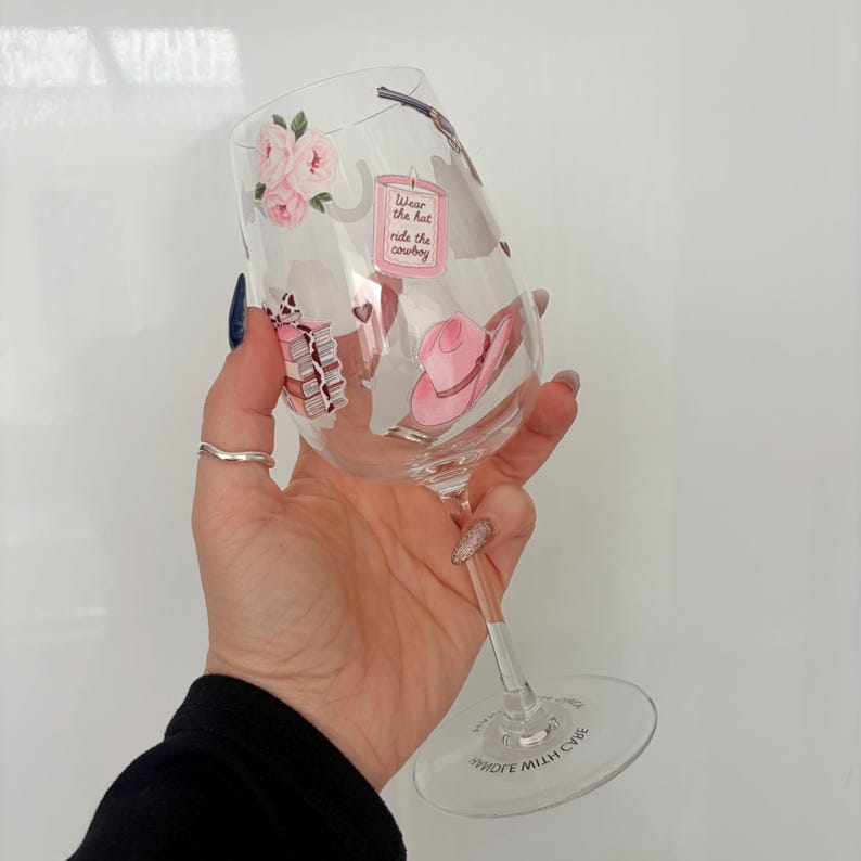 May include: A clear wine glass with a stem, decorated with pink roses, a pink cowboy hat, and the text "Wear the hat ride the cowboy". The glass has a wide bowl and a clear base.