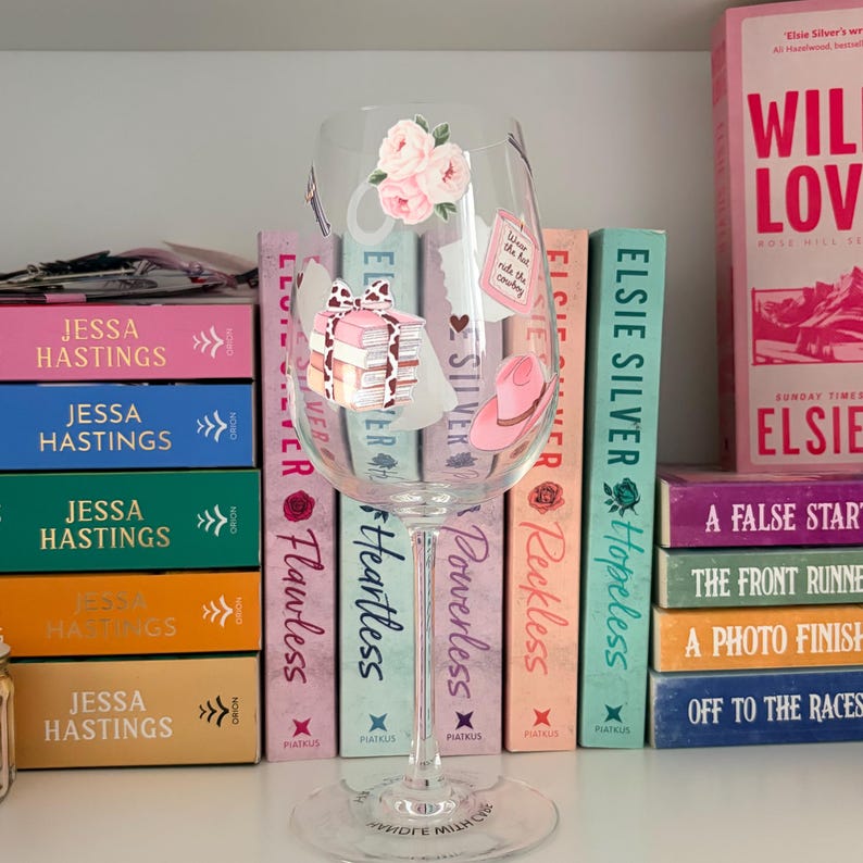 May include: A clear wine glass decorated with floral and book-themed stickers, standing in front of a shelf of books. The books have titles like "Flawless" and "Hopeless." The glass has a long stem and a wide bowl.