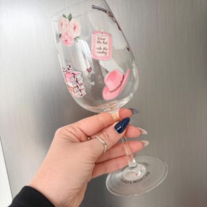 May include: A clear wine glass decorated with pink floral and cowboy-themed stickers. The stickers include a pink cowboy hat, a stack of books, and a candle with the text "Wear the hat ride the cowboy."