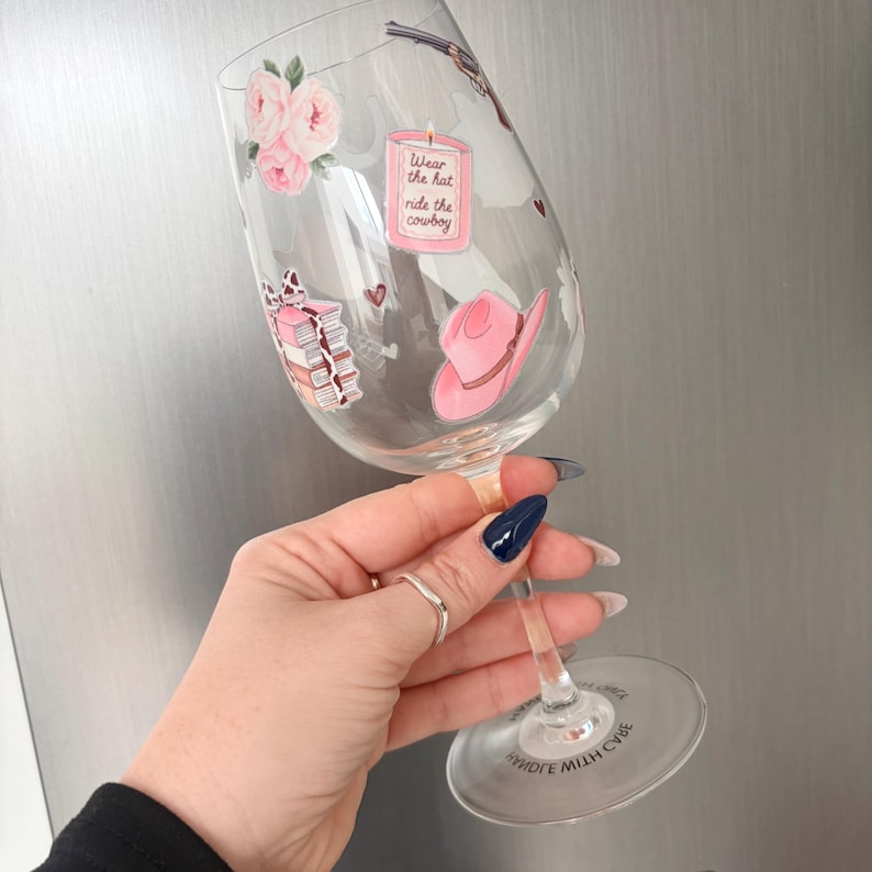 May include: A clear wine glass decorated with pink floral and cowboy-themed stickers. The stickers include a pink cowboy hat, a stack of books, and a candle with the text "Wear the hat ride the cowboy."