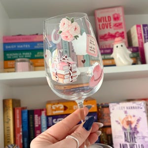 May include: A clear wine glass decorated with stickers of pink roses, a cowboy hat, and a stack of books tied with a cow-print ribbon. The glass has the text "Wear the hat, ride the cowboy". Bookshelves are in the background.
