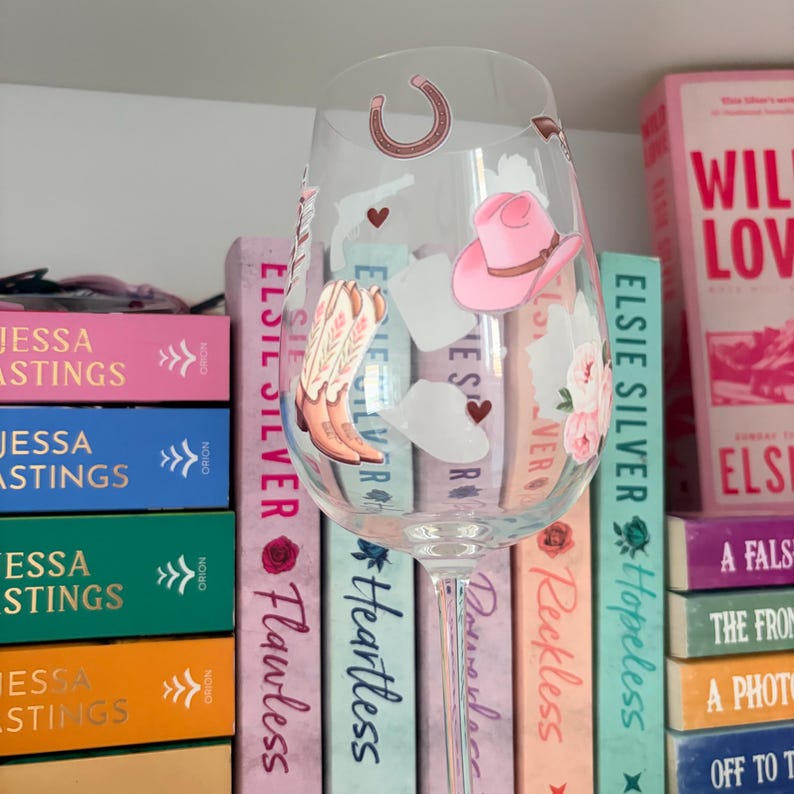 May include: A clear wine glass decorated with country-western themed stickers, including a pink cowboy hat, cowboy boots, and a horseshoe. The glass is in front of a shelf of books with pink, blue, green, and orange spines.