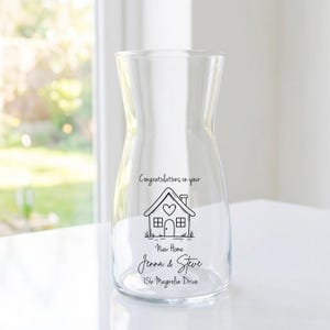 May include: A clear glass pitcher with a wide base and narrow neck. The pitcher has a black line drawing of a house with the text "Congratulations on your New Home Jenna & Steve 156 Magnolia Drive."