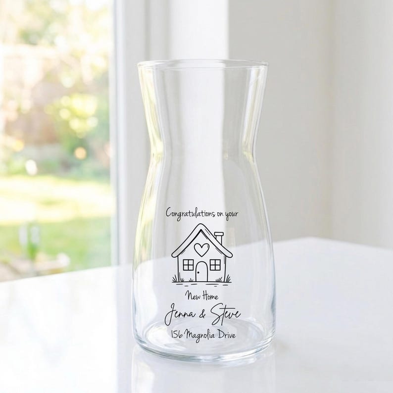 May include: A clear glass pitcher with a wide base and narrow neck. The pitcher has a black line drawing of a house with the text "Congratulations on your New Home Jenna & Steve 156 Magnolia Drive."