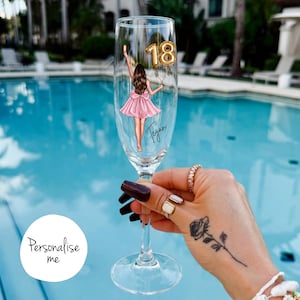 May include: A clear champagne flute with a custom illustration of a woman in a pink dress holding gold number 18 balloons. The name "Tegan" is written on the glass. The words "Personalise me" are in a white circle.