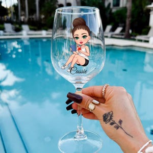 May include: A clear wine glass with a cartoon illustration of a woman reading a book, with the name "Cassie" below. The woman is wearing a white vest and black shorts. The glass is held up against a blue swimming pool.