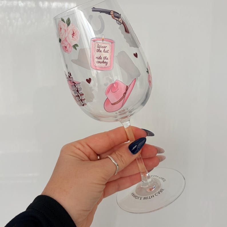 May include: A clear wine glass decorated with cowboy-themed illustrations. The glass features a pink cowboy hat, a gun, a horseshoe, and the text "Wear the hat ride the cowboy". Pink roses and hearts are also included in the design.