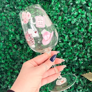 May include: A clear wine glass decorated with pink cowboy-themed stickers, including a pink cowboy hat, roses, and the text "Wear the hat, ride the cowboy." The glass is held against a green background.