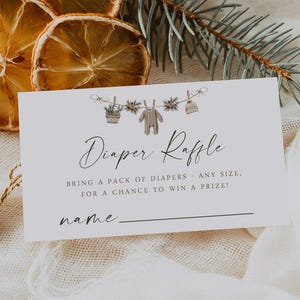May include: A baby shower diaper raffle card with the text "Diaper Raffle" and instructions. The card is decorated with a clothesline of baby items. Dried orange slices and greenery are arranged around the card.