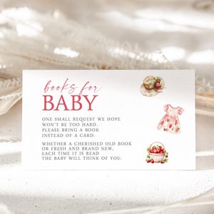 May include: A white card with red text that reads "books for BABY". The card has a watercolor illustration of a hat, a dress, and a basket of strawberries. The text on the card reads "One small request we hope won't be too hard. Please bring a book instead of a card. Whether a cherished old book or fresh and brand new. Each time it is read the baby will think of you."