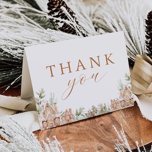 May include: A white card with the words "Thank You" written in brown script. The card is decorated with a watercolor illustration of a winter village scene.