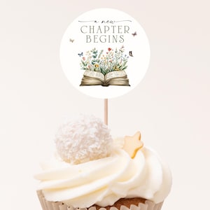 May include: A cupcake with white frosting, a coconut ball, and a star-shaped decoration. A round topper with the text "a new CHAPTER BEGINS" and a watercolor illustration of an open book with flowers and butterflies is on top.