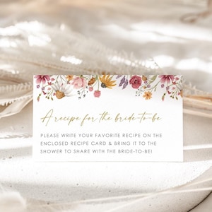 May include: A white card with a floral border and gold text that reads "A recipe for the bride-to-be". The text below the floral border reads "Please write your favorite recipe on the enclosed recipe card & bring it to the shower to share with the bride-to-be!"