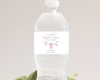 May include: A clear plastic water bottle with a white label that says "Sweet Baby Girl On Her Way" with pink and white baby clothes and a floral design. The label also includes the text "Amelia's Baby Shower".