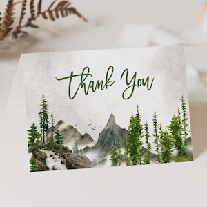 May include: A folded card with a watercolor landscape scene featuring mountains, trees, and a waterfall. The words "Thank You" are written in green script across the top. The card has a soft, neutral background.