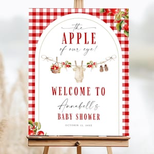 Peut inclure: Panneau de bienvenue pour une baby shower sur un chevalet en bois. Le panneau présente une bordure vichy rouge et blanche et le texte "The Apple of our eye !" avec "Welcome to Annabell's Baby Shower" en dessous.