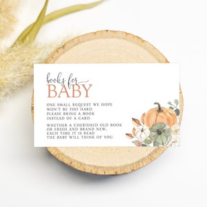 May include: A white card with the text "books for baby" in brown and black lettering. The card has a watercolour illustration of a pumpkin and flowers in shades of orange, green, and white. The text on the card reads: "One small request we hope won't be too hard. Please bring a book instead of a card. Whether a cherished old book or fresh and brand new, each time it is read the baby will think of you."