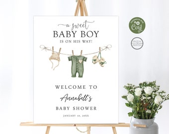 May include: A white sign with green text announcing a baby shower. The sign reads "a sweet BABY BOY is on his way!" and "WELCOME TO Annabell's BABY SHOWER JANUARY 10, 20XX". The sign features a watercolor illustration of a baby outfit, hat, and booties hanging on a clothesline.