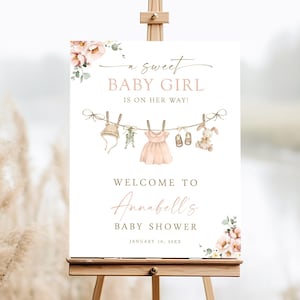 Può includere: Un cartello di benvenuto bianco con accenti floreali rosa e una corda di vestiti per bambini appesi a mollette. Il testo recita "a sweet BABY GIRL is on her way!" e "WELCOME TO Annabell's BABY SHOWER JANUARY 10, 20XX".