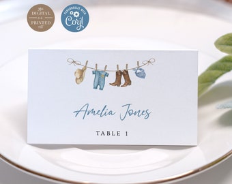 May include: A blue and white baby shower place card with a clothesline illustration featuring a baby outfit, cowboy boots, and a hat. The text "Amelia Jones" and "Table 1" are printed on the card.