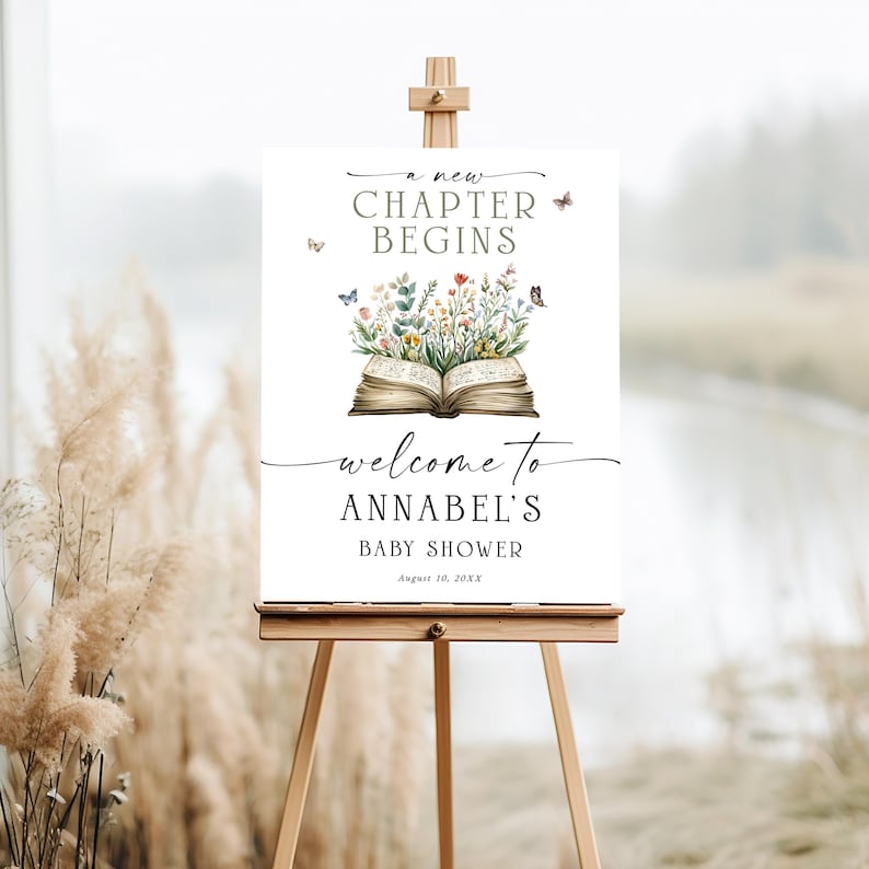 May include: A white welcome sign on a wooden easel, featuring the text "a new CHAPTER BEGINS" and "welcome to ANNABEL'S BABY SHOWER". The sign includes an illustration of an open book with flowers and butterflies.