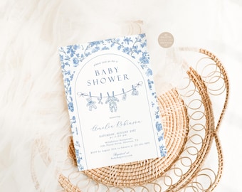 Puede incluir: Una tarjeta de invitación rectangular para baby shower con un borde de patrón floral azul y blanco. El centro muestra un marco arqueado con el texto BABY SHOWER sobre una ilustración de tendedero con artículos de bebé y detalles.