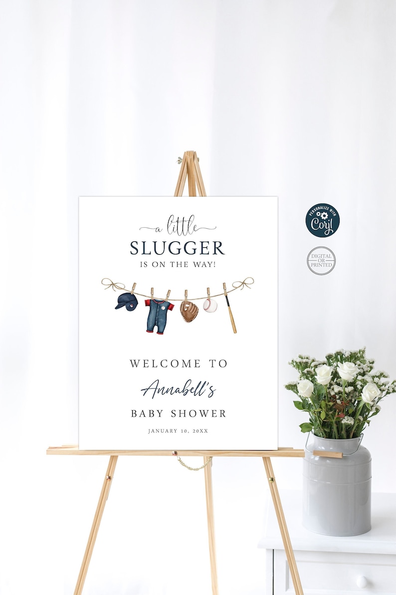 Peut inclure: Une pancarte blanche avec du texte bleu annon&ccedil;ant une baby shower pour Annabell. La pancarte indique "a little slugger is on the way!" et pr&eacute;sente une illustration sur le th&egrave;me du baseball d'une grenouill&egrave;re de b&eacute;b&eacute;, d'une casquette de baseball, d'un gant, d'une balle et d'une batte suspendues &agrave; une corde &agrave; linge.
