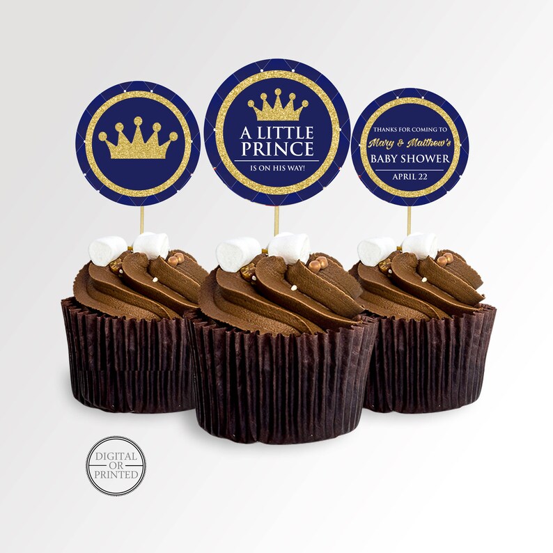 May include: Three cupcakes with chocolate frosting, topped with marshmallows and decorative toppers. The toppers are navy blue with gold accents and feature a crown and the text "A Little Prince". One topper says "Thanks for coming to Mary & Matthew's Baby Shower April 22".