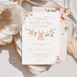 May include: A rectangular baby shower invitation card featuring watercolor floral accents and a clothesline illustration with a bonnet, dress, shoes, and a plush rabbit. The text reads A SWEET BABY GIRL IS ON HER WAY and includes event details.