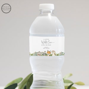 Op de afbeelding: Waterfleslabel met een witte achtergrond en een groene bladrand. Het label zegt "A Little Wild One Is On The Way!" met de tekst "Sarah's Baby Shower" eronder. In het midden van het label staan vijf bosdieren: een egel, een wasbeer, een vos, een uil en een hert.