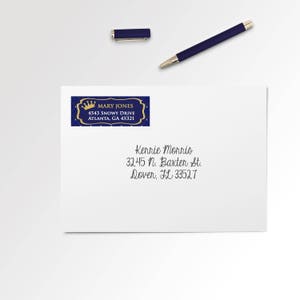 May include: A white envelope with a blue and gold address label featuring the name "MARY JONES" and address. A matching blue pen and pen cap are placed above the envelope. The envelope also has a handwritten address.