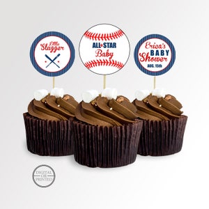 May include: Three chocolate cupcakes with baseball-themed toppers. The toppers feature baseballs and text that reads "Little Slugger", "All-Star Baby", and "Erica's Baby Shower Aug. 15th". Each cupcake is topped with a marshmallow.