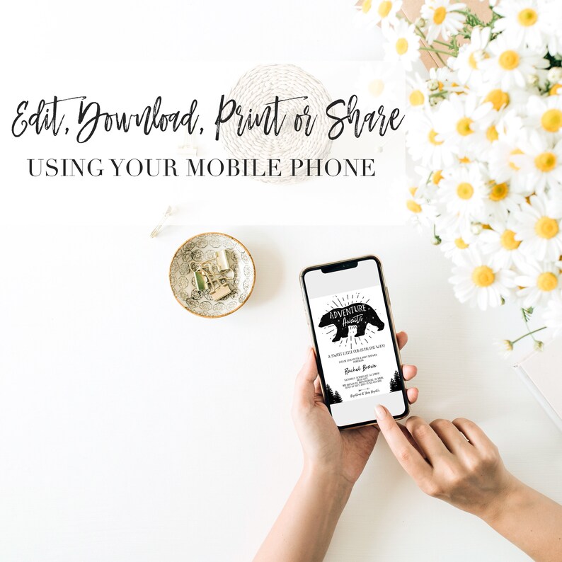 May include: A smartphone displaying a printable invitation for an "Adventure Awaits" party. The invitation features a black silhouette of a bear with the text "Adventure Awaits" in a circle above the bear. The text "A baby shower celebrating the arrival of our little adventurer" is below the bear. The invitation is on a white background.