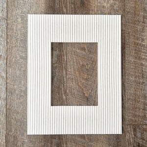 May include: A rectangular picture frame with a white and gray striped border. The center of the frame reveals a wooden surface. The frame is set against a wooden background.