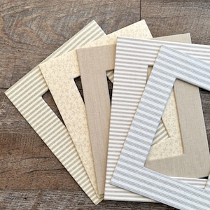 Può includere: Una collezione di cornici rettangolari con vari motivi in tessuto. Le cornici presentano strisce, stampe floreali e colori neutri a tinta unita come beige e crema. Le cornici sono disposte su una superficie di legno.