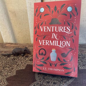 Op de afbeelding: Een boek getiteld "Ventures in Vermilion" met een rode kaft en decoratieve illustraties. De kaft bevat een vergrootglas, een hoed en andere detective-thema-elementen. De naam van de auteur, J.T. Thompson, staat onderaan gedrukt.